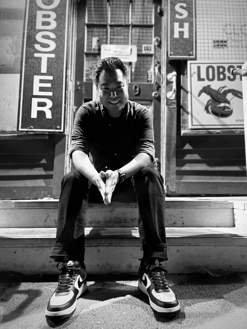 A black and white picture of a man wearing a cuffed shirt, black pants and sneakers sitting down on the steps of a building with signs for lobster outside.