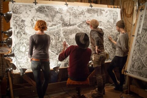 Four illustrators stand facing a large wall-sized drawing of Mesoamerica Resiste, a poster made by 23 anonymous volunteers and 10 years of time