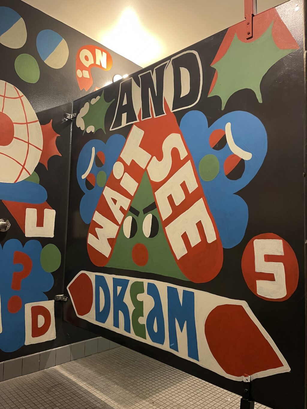 A text based blue, red, green, black and white text bubble mural in the SPACE bathroom that says "Wait and See" and "Dream" 