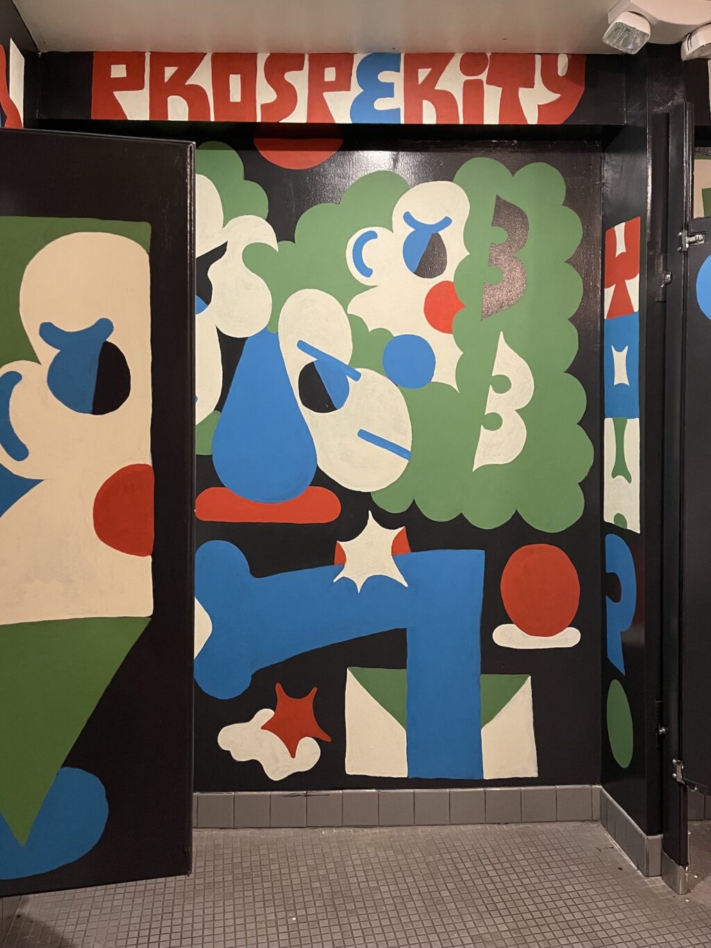 A mural in the SPACE restroom in blue, green, white, red, and black that is graphic shapes, cloud bursts, and faces with the word "Prosperity" written overhead. 