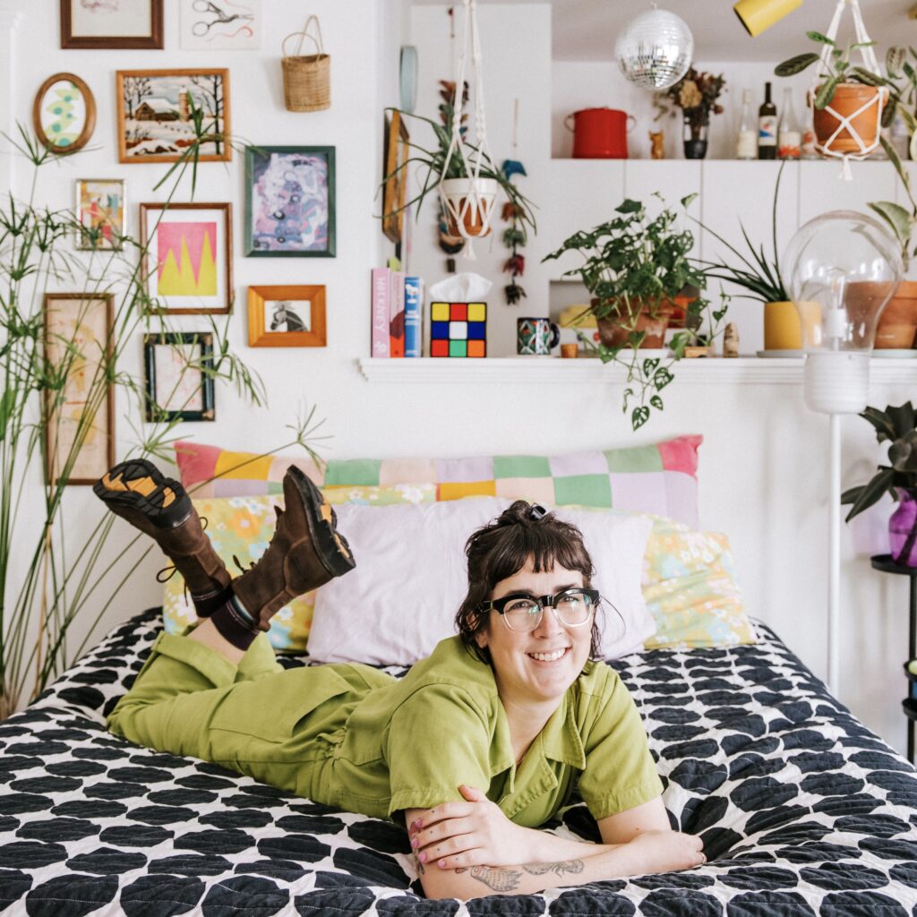 Hannah Hermes wears a lime green jumper and hiking boots laying on he torso on her bed, with a panoply of interesting art and horticulture hanging on the wall behind her