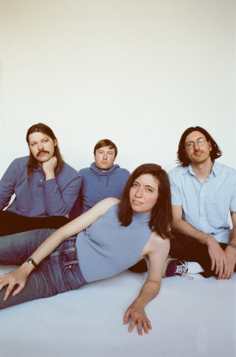 Ratboys posed against a white background, sitting and lying on the ground, all wearing light blue shirts, sweaters, and sweatshirts with dark blue and black pants.