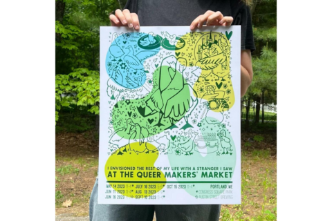 The artist holds up a large colorful poster for the Queer Makers' Market