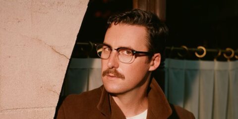 Nick Waterhouse in a brown overcoat and horn rimmed glasses looking into the camera.