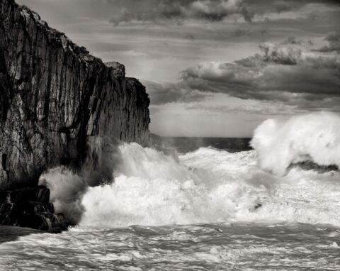 A black and white photograph of white foamy waves crashing against rocks by Carl Austin Hyatt