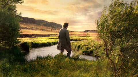 A man in a long trench coat stands facing away from the camera as he looks over a vast expanse with mountains, grasslands, and a river under a beautiful sky