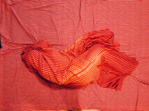 A form that appears to be a body wrapped in a red striped fabric, laying on the same red fabric.