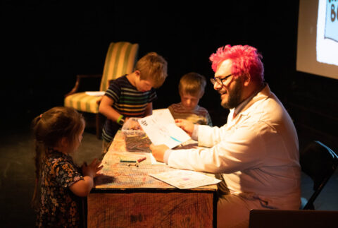 Ricky Rainbow Beard, a nonbinary person with red dyed hair in a white lab coat, sits at a desk while kids peruse the wares