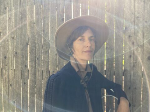 Josephine Foster, a white woman wearing a brimmed straw hat, smiles at the camera from the inside of what appears to be a barn, with light flares hitting the lens