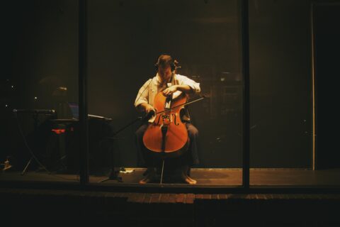 Robin Lane, a white man with a white button down shirt and black pants, plays a cello while seated inside SPACE's Window Gallery enclosure