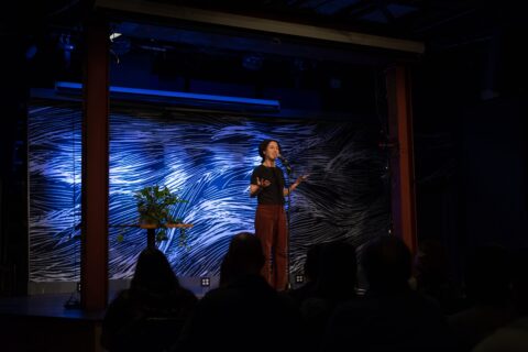 A woman with a black shirt and red pants stands on the blue-lit SPACE stage with her palms up, telling a story, while a plant sits on a side table