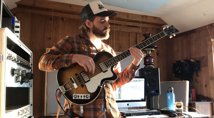 Hamilton Belk in a recording studio, playing a Beatles-style bass guitar in a plaid shirt and trucker hat. 