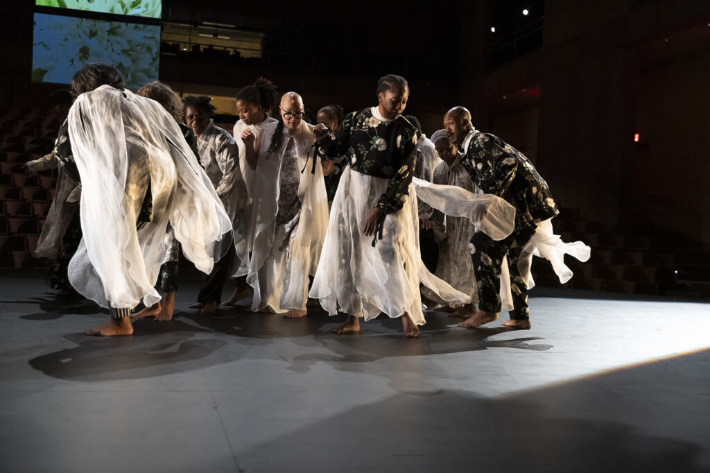 A dozen members of Scapegoat Garden dancing together in a tight group, with flowing white and black floral fabrics, each costume unique to each dancer.