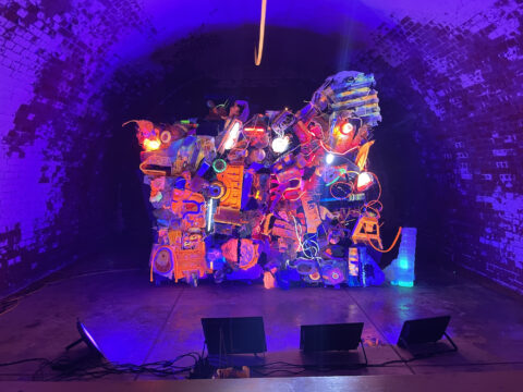 An image of a monumental sculpture made up of a pile of smaller parts in black light, much of it glowing orange.