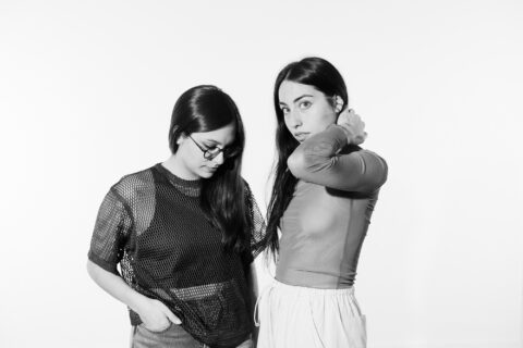A black and white photo of Deerlady's Mali Obomsawin and Magdalena Abrego standing together in studio light. Mali is touching her hair and looking over her arm at the camera. Magdalena is looking down at the ground with a hand in her pocket.