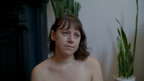 Joanna Arnow, a white woman with shoulder length brown hair, is seen shirtless with an expressionless face inside a plain-looking domestic room with white walls