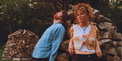 Both members of Babehoven leaning against a rock. Maya is wearing glasses, a fringed colorful vest, and looking down at the ground. Ryan is wearing a blue sweatshirt and staring up at the sky. They look great.