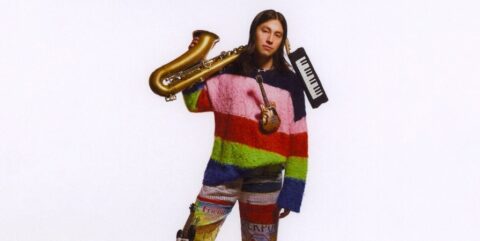 Sen Morimoto in colorful striped sweater, saxophone over one shoulder, keyboard on the other shoulder, and mini guitar hanging around his neck.