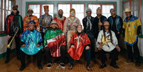 The modern day Sun Ra Arkestra led by Marshall Allen. All 13 members are dressed in colorful outfits. Five sit in front. Eight stand in the back.