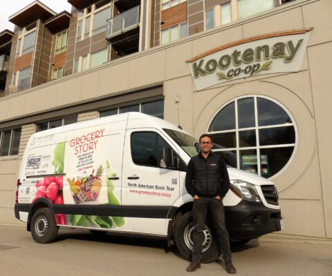 Jon Steinman, a white man, stands next to a co-op and a van advertising his book
