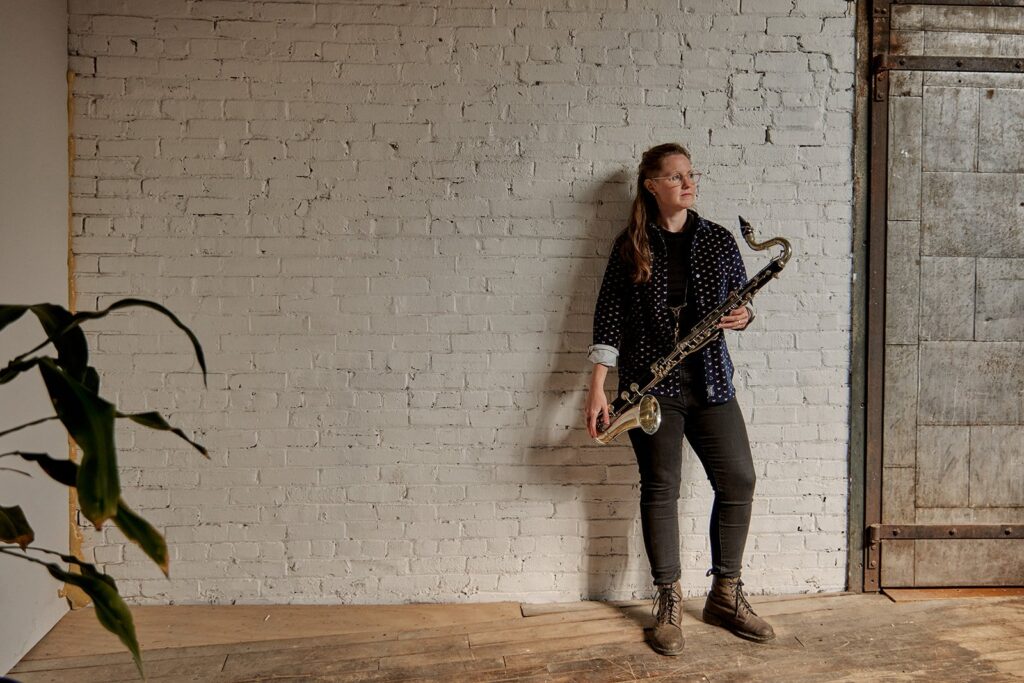 Allison Burik holding a bass clarinet standing against a brick wall painted white. A house plant infiltrates the corner of the frame. 