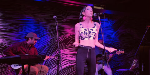A photo from the 2023 Lifesongs performance at SPACE. A singer with a bandana on their hair, blue eyeshadow, and red earrings sings in front of the mic with a guitarist and bassist behind them.