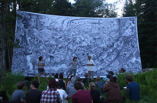 Three artists from the Beehive Collective stand speaking in front of a gigantic print— at least 12 feet high and 20 feet wide. It's hung between trees outside and an audience of sits on the grass in front. 