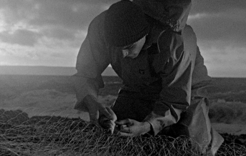 A black and white grainy still of a maritime worker cutting rope.