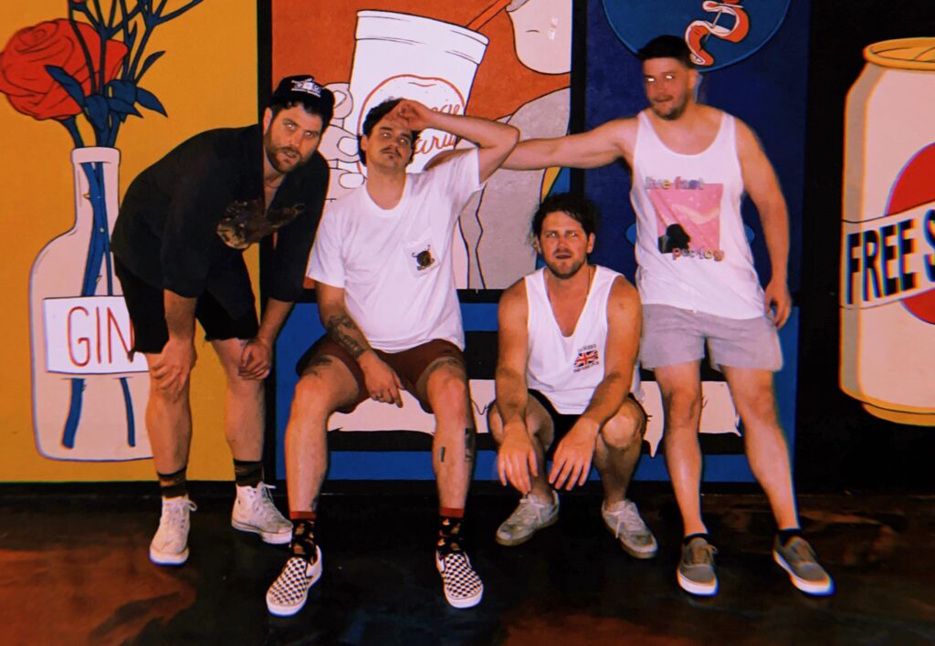 The boys of Greasy Grass looking ready for summer, sporting tank tops, shorts, and sneakers, in front of a colorful mural wall.