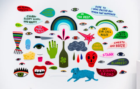 A series of shaped paintings on wood panels mounted on a white wall including a mouth, blue dog, wine bottle, hand, red skull, and a series of talk bubbles with various statements.