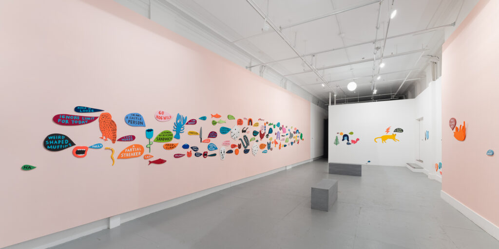 A gallery installation image of the pink and white walls with salon style hangs of numerous colored talk bubble shapes and colorful animals by Martha Rich. 