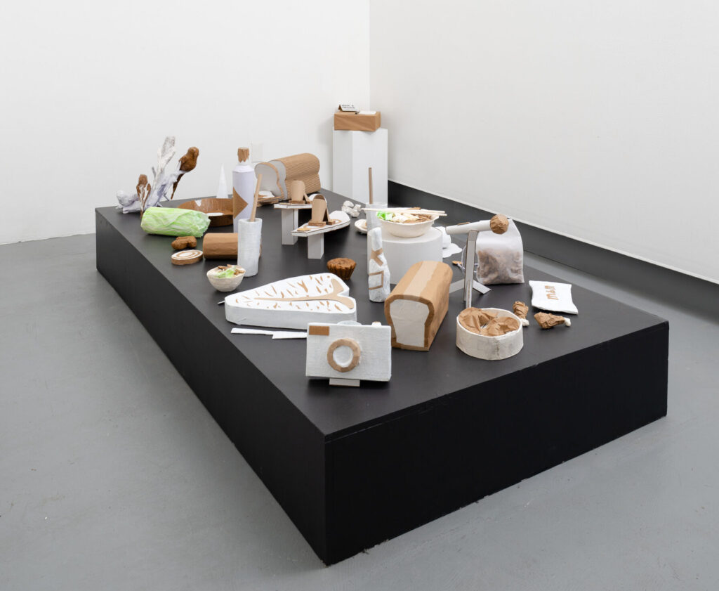 A black pedestal with a series of paper an cardboard sculptures depicting meals and objects that are personal.