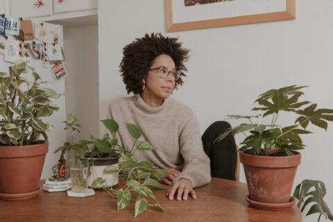 Ayana Elizabeth Johnson, a young Black woman with curly hair, sits at a desk in an office with several potted plants. She looks to the side off camera.