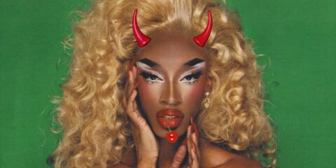 A close-up of drag queen Lana Jarae with devil horns and a cherry hanging from her mouth. She's set against a green background.