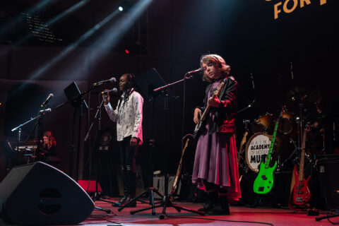 two pre-teen kids are seen on stage playing music, one singing into a microphone and the other playing a bass guitar