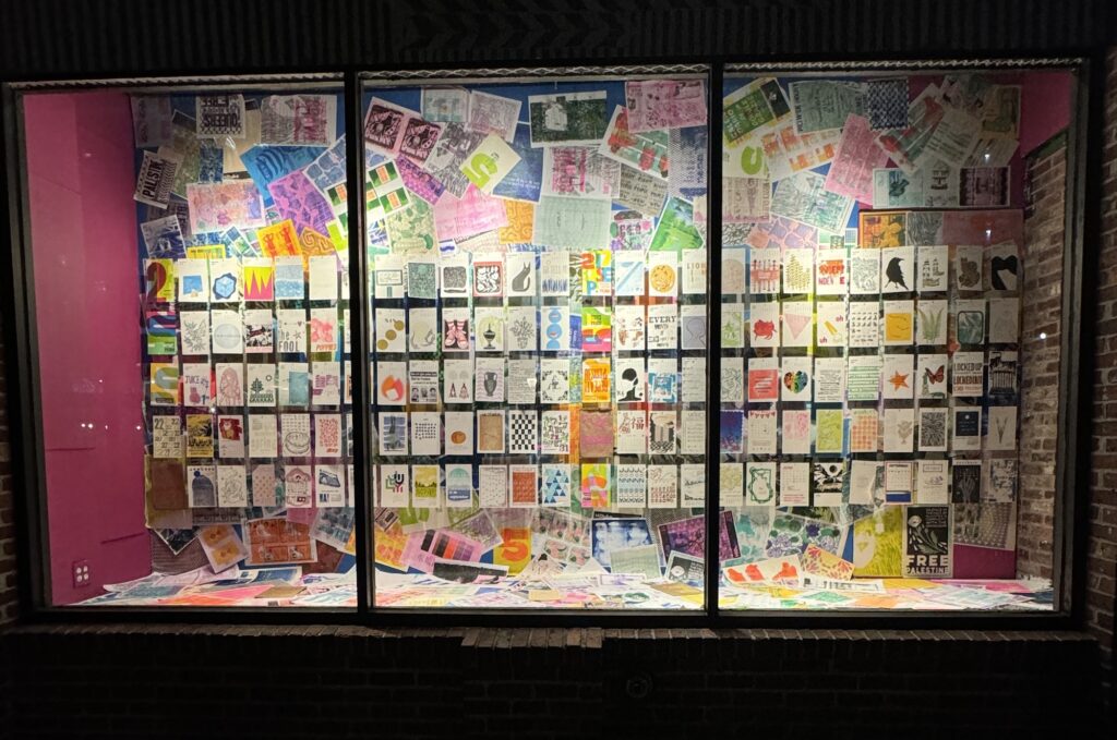 A photograph at night of the glowing Pickwick Press window installation featuring 10 years of their calendar art, with a wheat pasted wallpaper of test prints behind it.