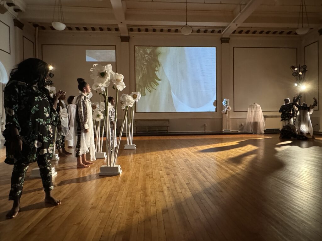 On the left three figures are looking right surrounded by white human height flower props, on the right, three figures are caught mid-dance with a white light behind them in silhouette. 