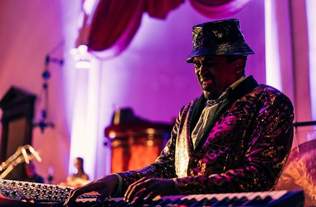 An image of a member of Sun Ra Arkestra playing keyboard passionately in a shiny costume and hat with a purple background. 