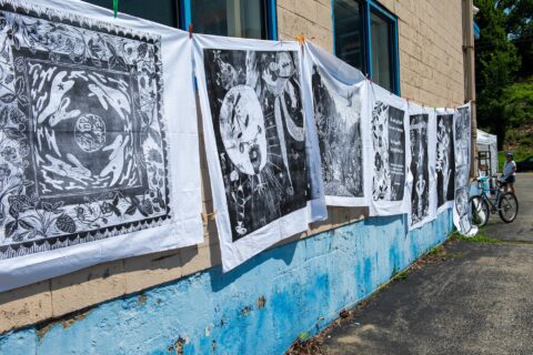 Large scale woodcarved prints of black ink on white sheets hanging along the side wall of Running With Scissors outside in the sunshine on a clothes line.