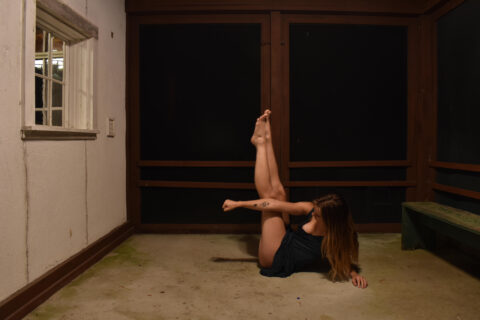 a young dancer in a dark lit room with long wavy brown hair and a black leotard balances in a cross-body position with legs pointed up in the air