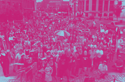 A pink and blue stylized photo of a crowd of Dyke March Maine participants in Congress Square Park smiling at the camera.