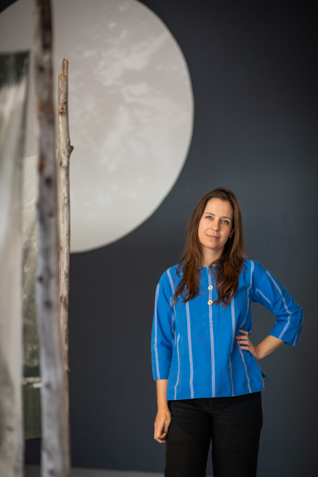 Iris Williamson, a white woman with shoulder-length black hair wearing a blue shirt with white stripes stands with her arm on her hip while looking at the camera. She stands before a work of art in the background that resembles a large moon, with half of it visible in the frame.