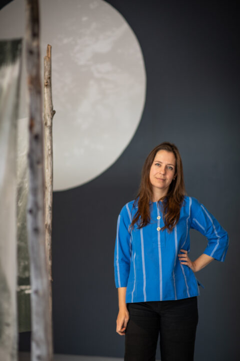 Iris Williamson, a white woman with shoulder-length black hair wearing a blue shirt with white stripes stands with her arm on her hip while looking at the camera. She stands before a work of art in the background that resembles a large moon, with half of it visible in the frame.