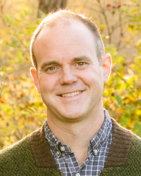 Zach Hefferen, a white man wearing a blue and white collared shirt, stands smiling at the camera before a backdrop of trees with a light yellow fall foliage