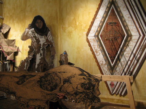 An installation shot of a wheat pasted skull, woman in a head covering, and diamond shaped decorative pattern against a yellow backdrop.