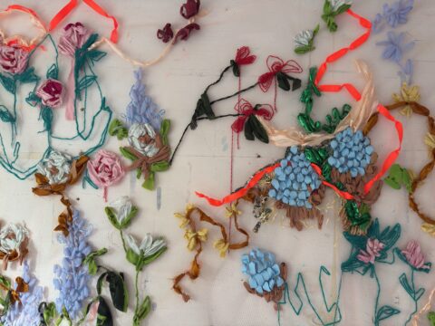 In this work by Phoebe Harris, a collection of materials arranged in floral patterns are affixed to a mesh netting on a wall