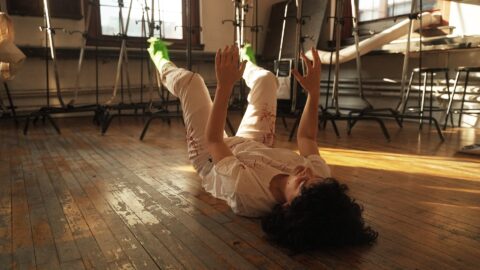 A film still of the artist, Martha Schnee, a white woman with dark curly hair, on a rough worn floor in a sun beam with her hands and legs up in the air with costumed frog legs on her feet.