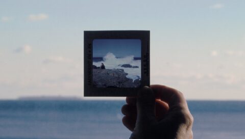 A still from Erin Johnson's film, a hand holds an old square piece of slide film of waves crashing on the coast with two figures on the rocky shore.