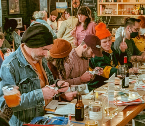 A group of people in various colored hats sit at a table with scissors making collages.