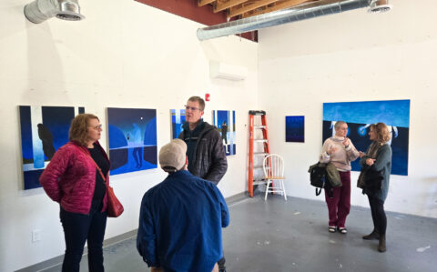 A group of people in front of blue paintings with abstracted figures in the landscape in a white walled art studio setting.